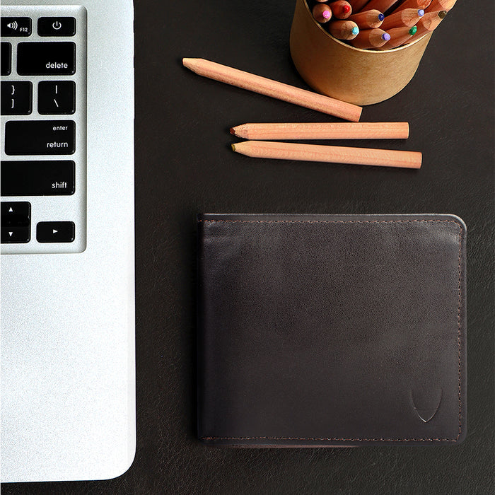 Brown leather bi-fold wallet beside laptop and pencils, featuring embossed Hidesign logo and crafts.