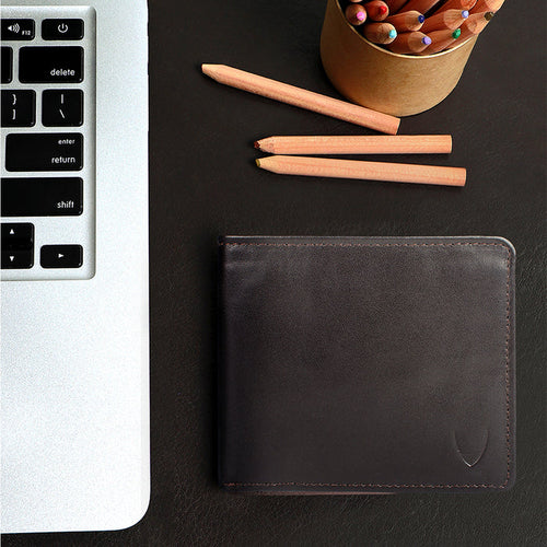 Brown leather bi-fold wallet beside laptop and pencils, featuring embossed Hidesign logo and crafts.