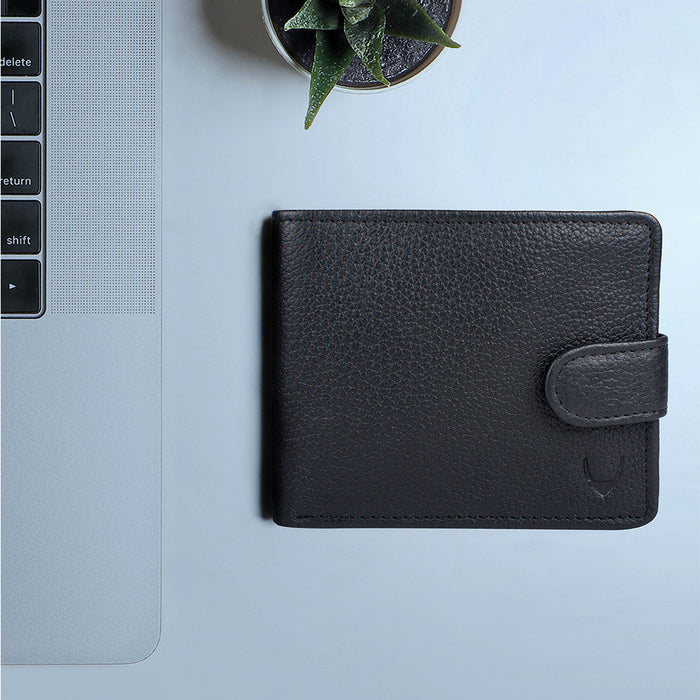 Black bi-fold leather wallet by Hidesign placed on a modern desk beside a laptop and a plant setup.