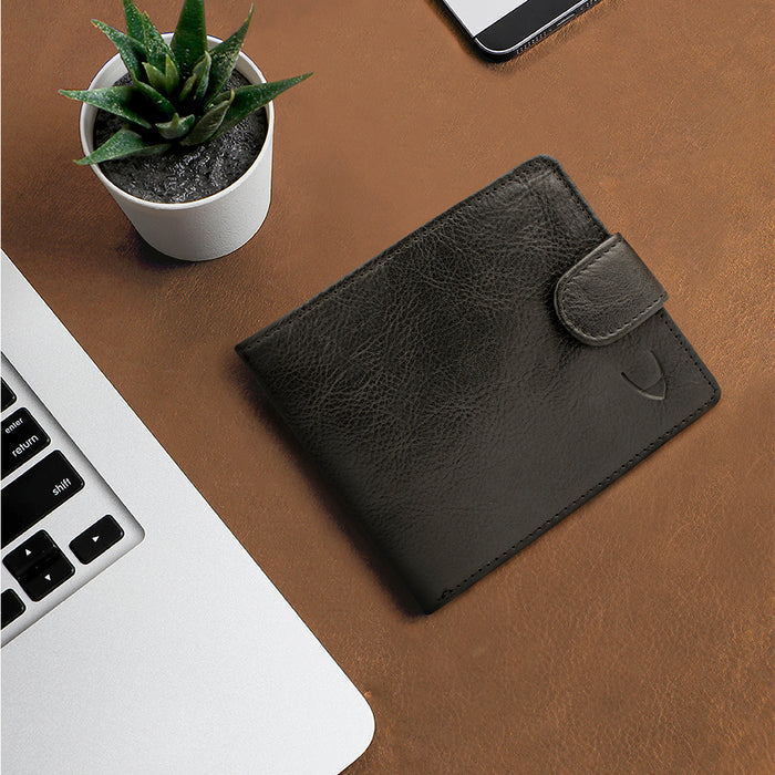Black leather bi-ranch fold wallet, placed on desk beside laptop and plant setup in the background.